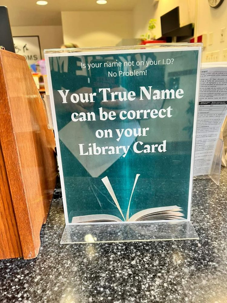 A sign in a library with a green background and white text. The text reads "is your name not on your I.D.? No problem! Your true name can be correct on your library card".

W librarians.