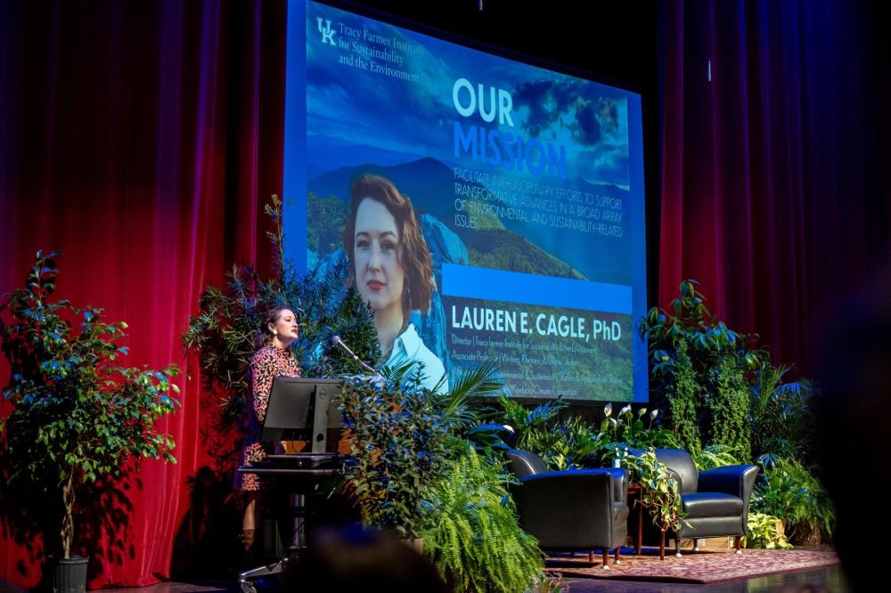 Photo of a white woman in a floral dress at a podium in front of a large projection of her headshot and surrounded by lush green foliage.