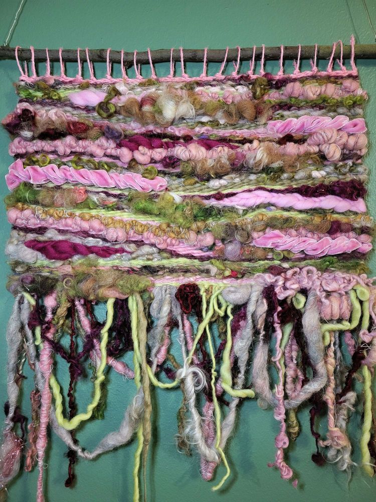 hand woven tapestry in shades of green, magenta, and pink, with multiple textures and density of fibers, hanging against a green wall.