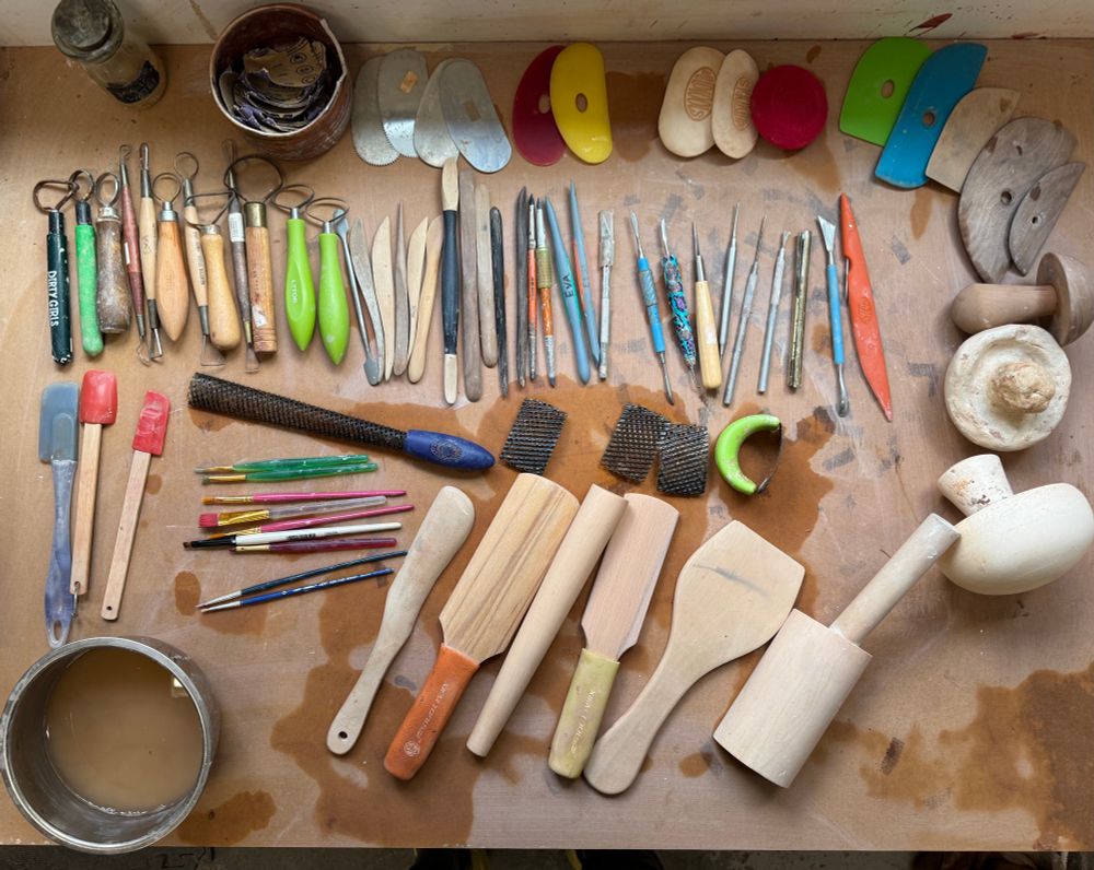 A whole desk of clay tools. Starting at the top: sand paper, four metal ribs, 4 rubber, 2 wooden, handful of sponges. 12 ribbon loop tools. 8 curved wooden tools, two of buffalo horn, 4 rubber smoothing tools. 5 bladed tools, a handful of needle tools. 3 mushroom shaped anvils out of wood or clay. 3 spatulas, 4 clay shredders/ surforms, 8-10 brushes, and 6 large wooden paddles.