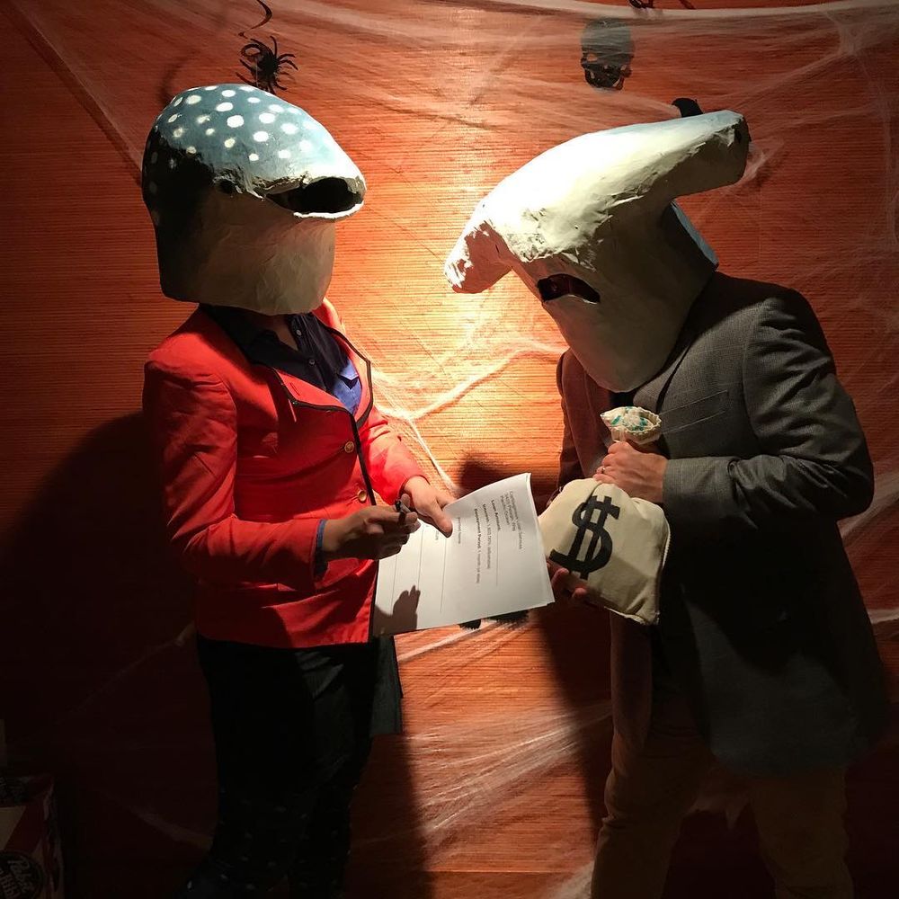 A person on the left with a paper mache whale shark mask. They’re holding a contract. On the right someone with a hammerhead mask who is wearing a poorly fitting suit, and holding a fake bag of money.  