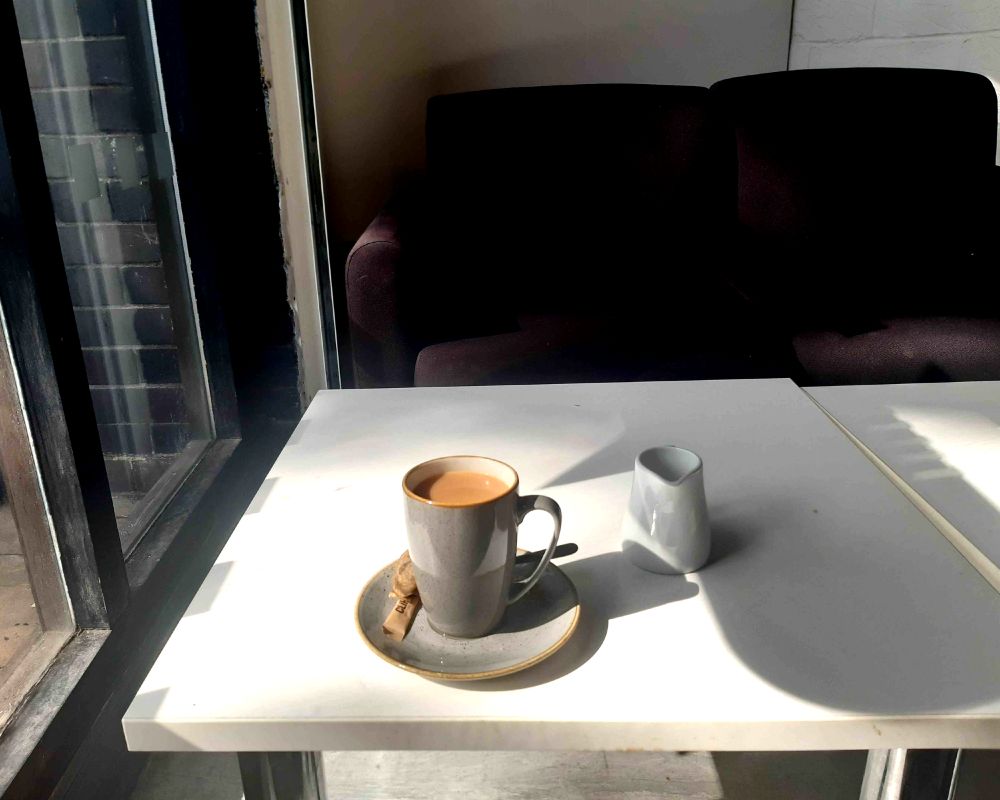 A photo of a white table, light falling onto it from the large window to the left. A sofa is in the background. On the table, within a white mug, is a cup of tea, alongside a milk jug.