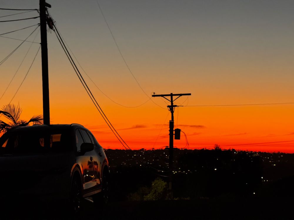 Black silhouettes of power lines agains an orange to gray blue gradient sunset. You can see reflection of the sunset off of a parked car