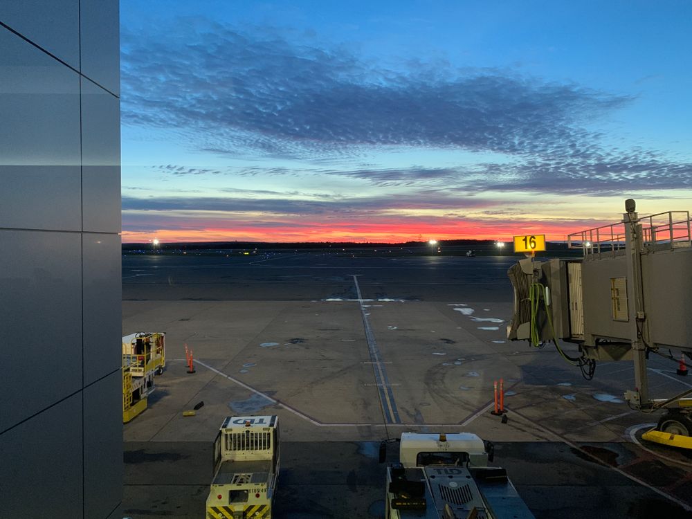 A sunrise at the airport with a jetway and service vehicles but no plane.. yet 