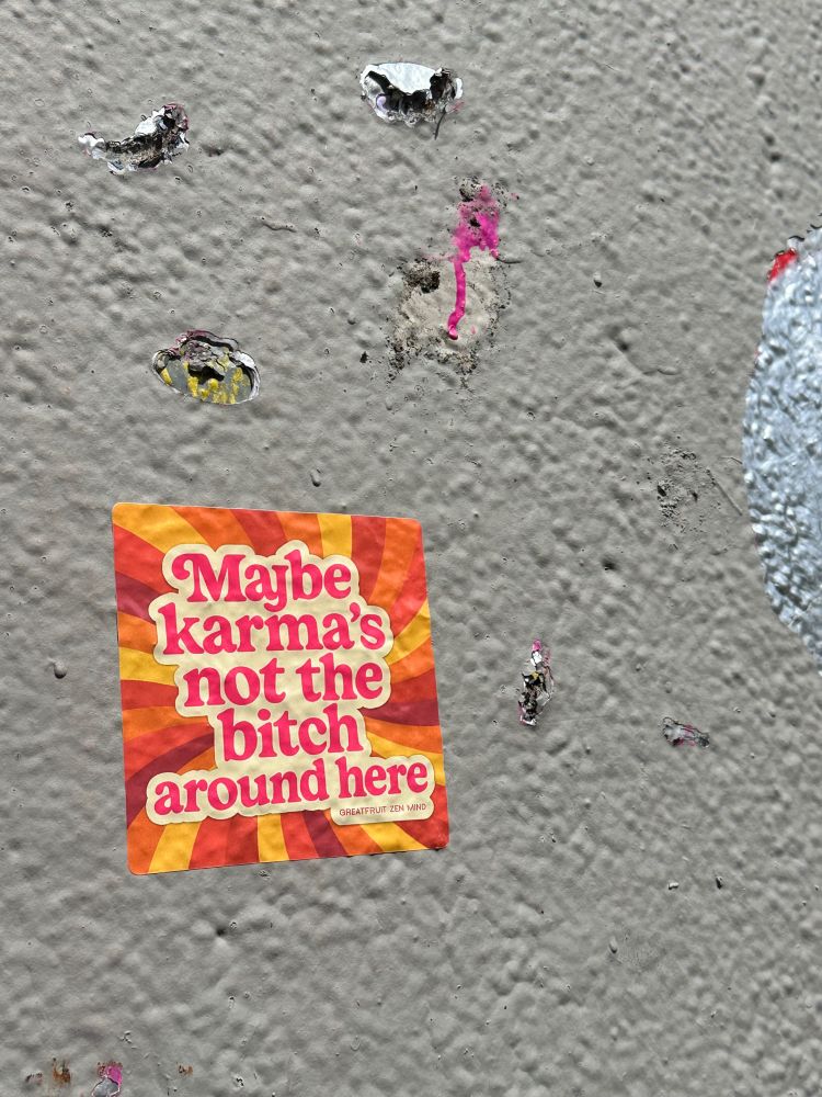 The "Maybe karma's not the bitch around here" sticker hung on an exterior concrete wall. Sticker contains text, against swirling lines of yellows and oranges.