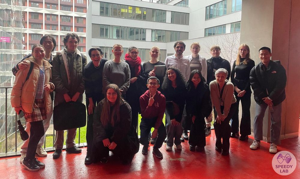 A group of people is standing in a university building, in front of a glass wall. They are smiling at the camera.