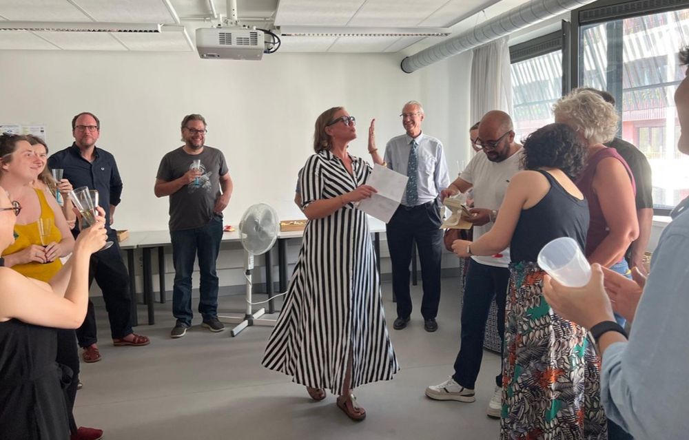 Anne Hermes is standing in a circle of smiling people. She is holding a sheet of paper in her hand and is blowing kisses.