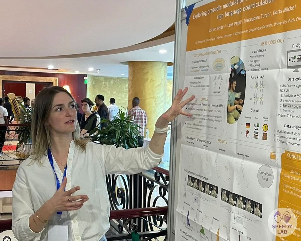 Justine is standing on the left-hand side of a poster that presents results on sign language coarticulation. She is in the middle of talking and is looking as well as gesturing towards the poster.