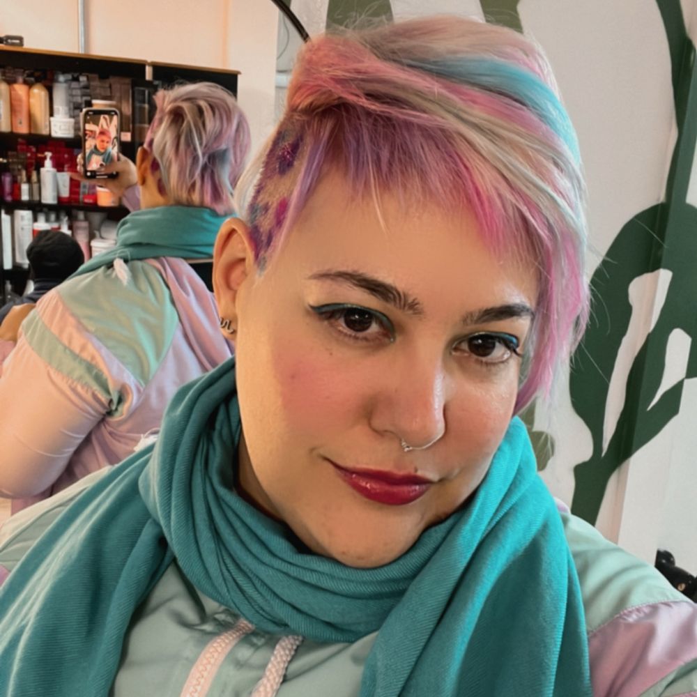 Selfie com meu cabelo platinado com mechas rosa, roxa e azul e a lateral raspada com oncinha colorida. Estou usando um delineador cromado verde e azul e um protetor labial rosado. Atrás de mim um reflexo do meu cabelo no espelho do salão