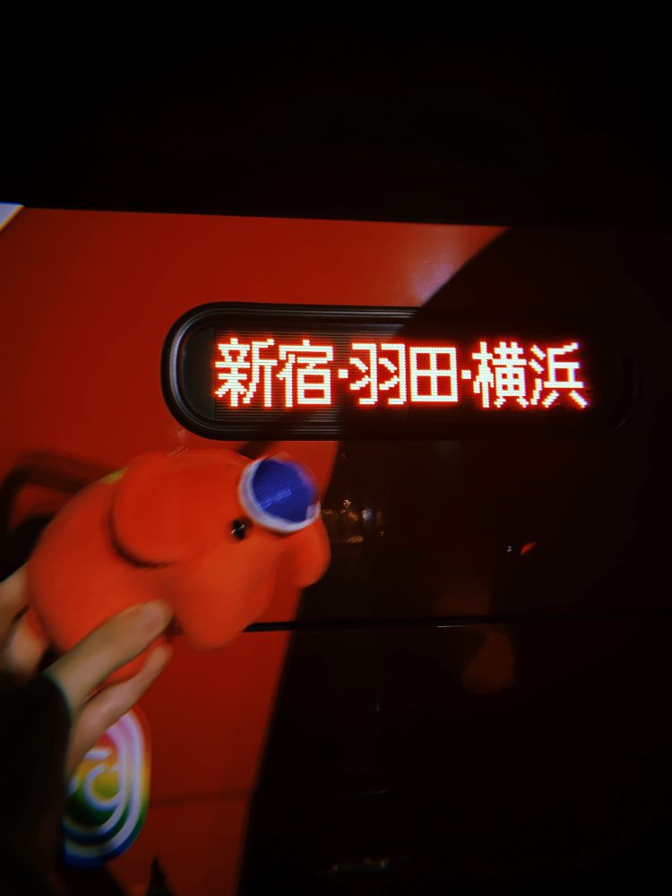 「新宿・羽田・横浜」の電光掲示板の前にアカレンジャーどむぞうくんを掲げている写真