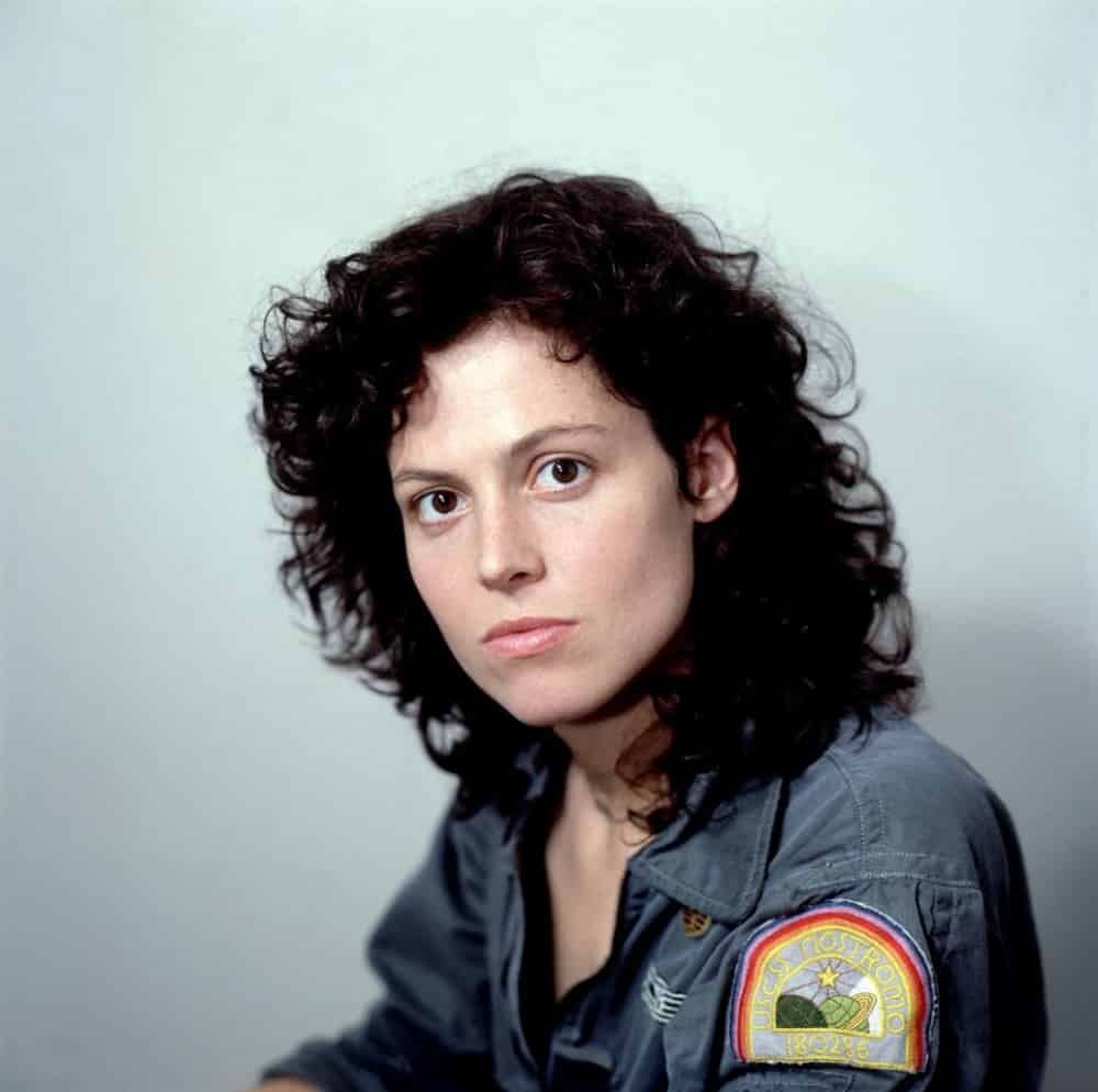 Lt. Ellen Ripley (played by sigourney weaver) of the nostromo mining vessel, she sits for a photo, wearing a blue jumpsuit with a nostromo patch on the shoulder. From alien 1979