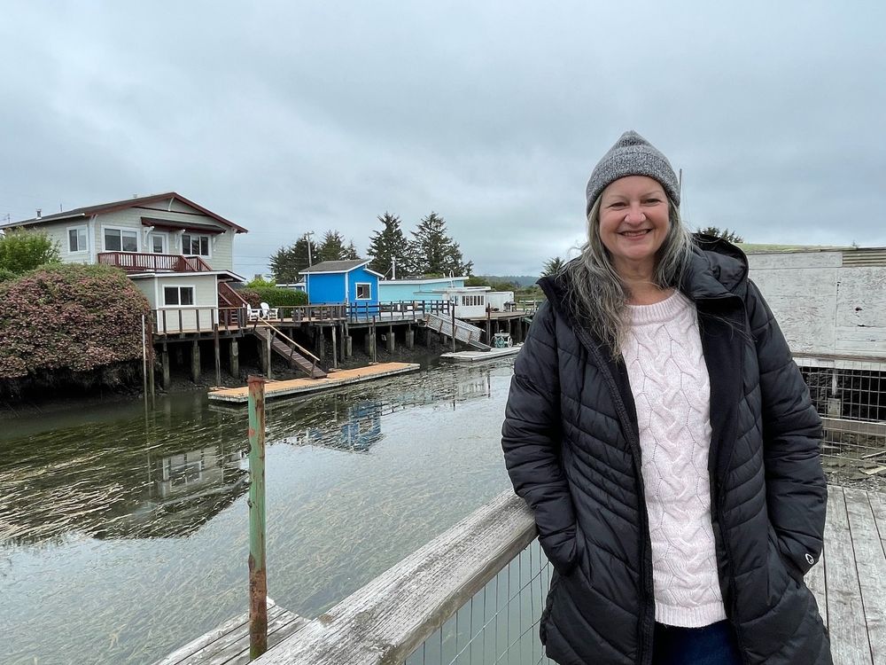 A smiling woman in a winter coat and hat stands on a deck next to a canal.