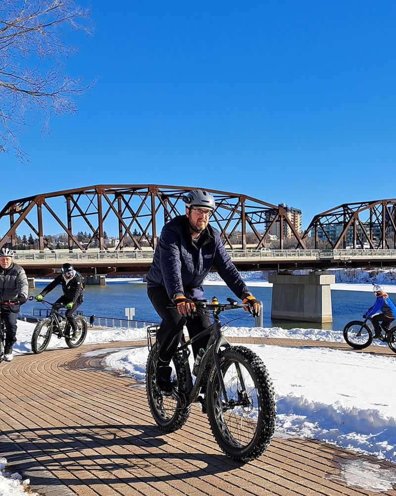 Winter cyclists riding up a brick path with the Victoria Bridge and South Saskatchewan River in the background, Saskatoon, SK. 