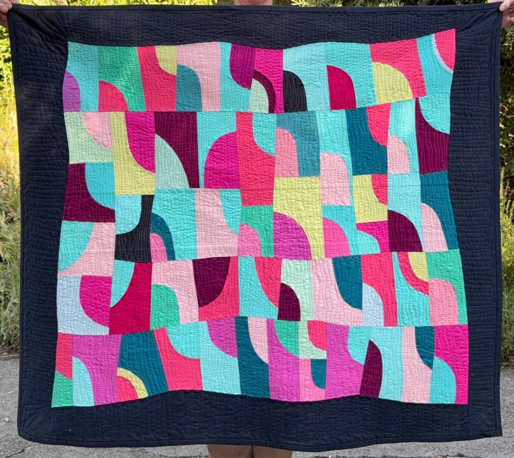 A quilt of green, blue, pink, and purple blocks with quarter circle, and quarter ovoid shapes, framed by darker fabric border that is wavy. Approximately 47 inches by 42 inches. 