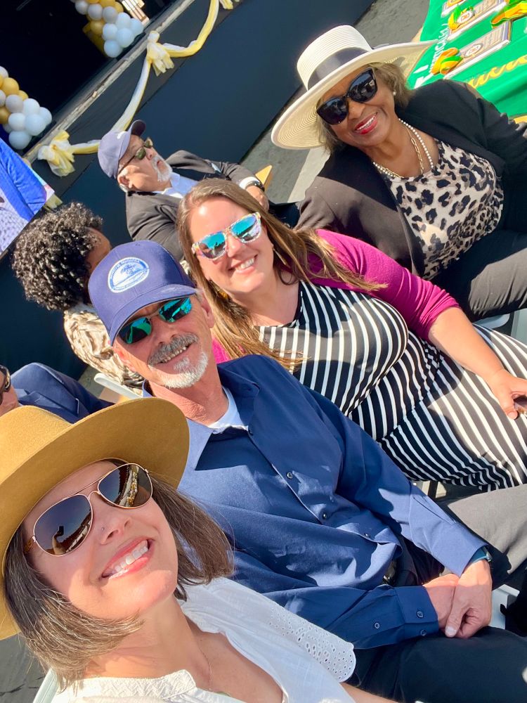 I woman takes a selfie with three other people looking at the phone. They are on a stage in bright sun, with some bunting in the background. 