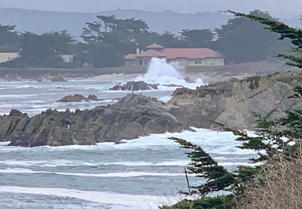 A landscape photo looking up a rocky coastline on a foggy morning. There is a large wave hitting the shore. A beige, single-story building with a clay tile roof sits in the background, a bit further up the coast. 