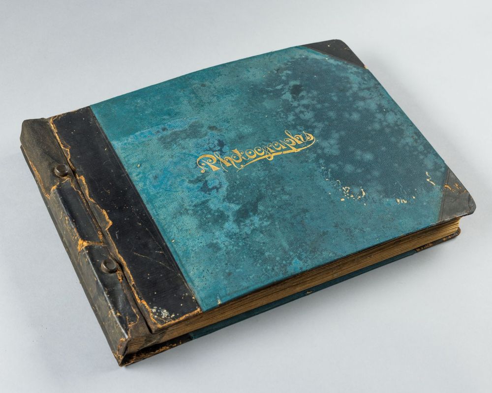 A photo of a very old looking album that has a waetherd black leather binding and a blue cover with the word "photographs" on it. 