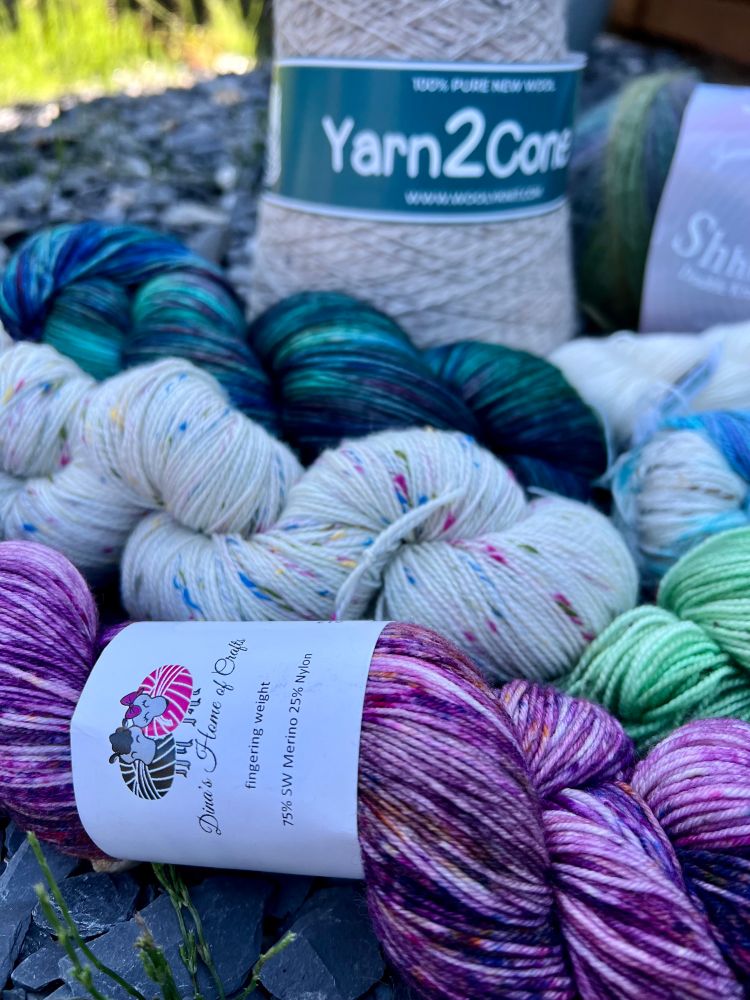 Three skeins of hand dyed yarn in purple, cream with rainbow spots, and deep blue/green. Behind them is a cone of 2ply wool yarn in natural colors. 