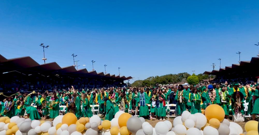 A view from a stage out to the main arena at Monterey County Fairgrounds. Hundreds of high school graduates in green cap and gown regalia are seen in front of the stage. 