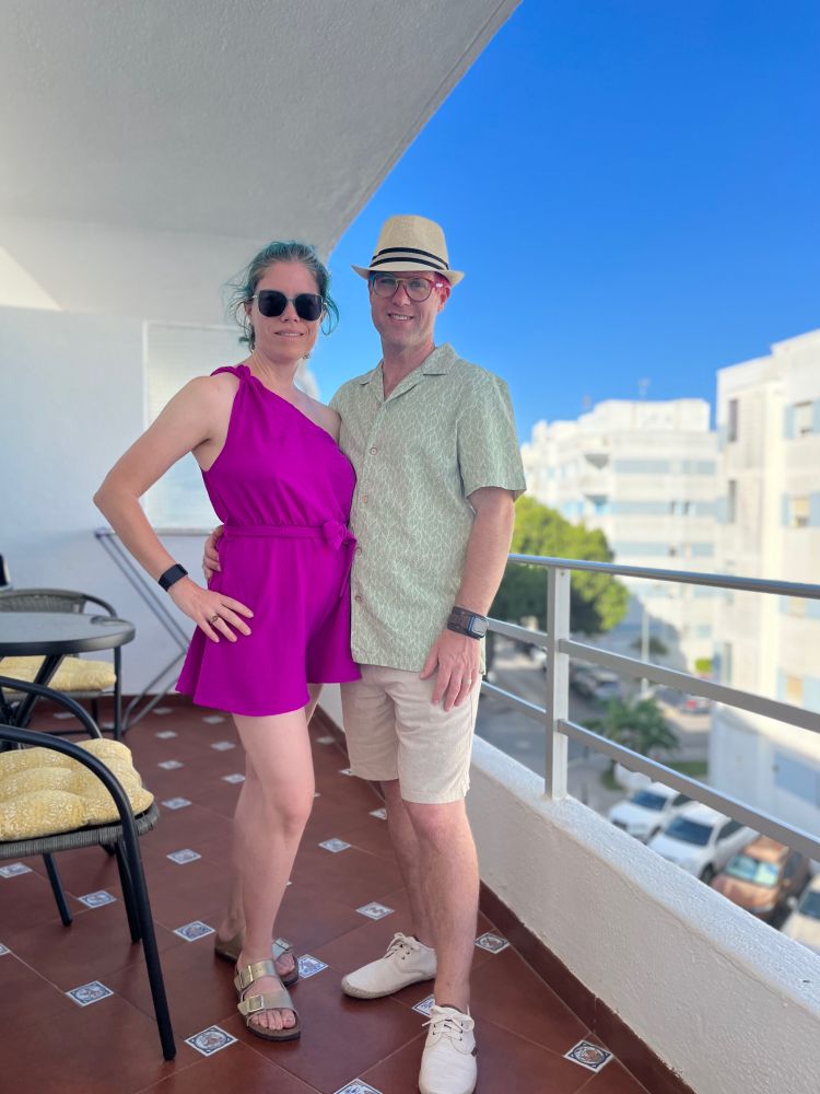 A man and a woman standing on a Spanish tiled balcony. She’s in a fuschia one shoulder romper with gold Birkenstocks. He’s wearing linen shorts and a green and brown linen shirt. 