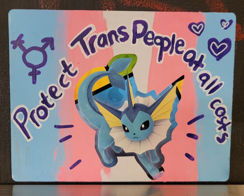Photo of sticker on a wall. Sticker has a painted trans pride flag in the background. The center is a vaporeon (pokemon). Text says, "Protect Trans People at all costs." Left corner has a trans symbol, right corner has a bunch of hearts. 
