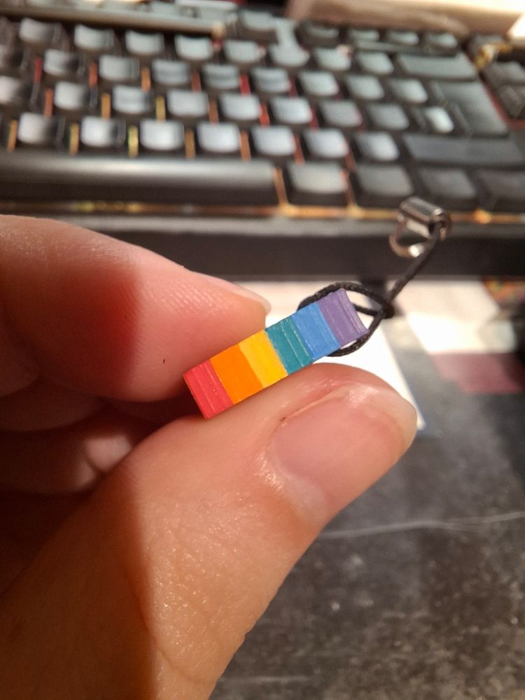 A close-up of a miniature brick painted with the rainbow flag, held between a thumb and a finger. 