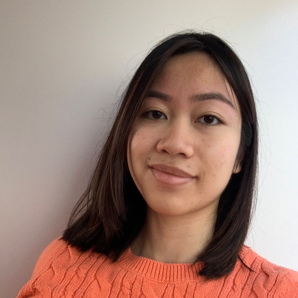 A headshot selfie of me against a white background. The subject is closed-mouth grinning at the camera wearing an orange-coral sweater, with straight black hair with dark brown highlights just below the shoulders and groomed, slightly arched brows.