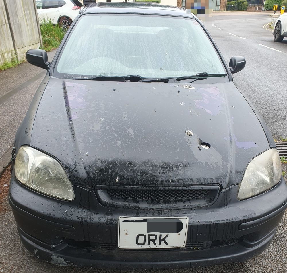A black car with 'ORK' in the numberplate; the rest of the numberplate has been pixilated for security purposes, as has a blue car in the background.
