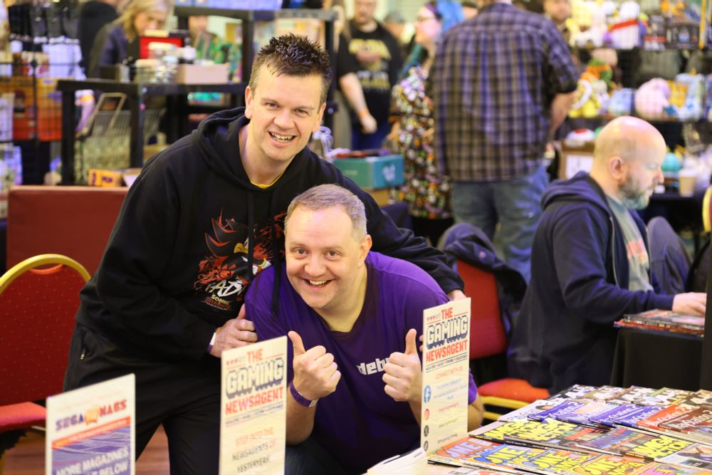 Marc Jowett and Dan Morris of Debug magazine, posing behind a table at Play Expo Blackpool 2025