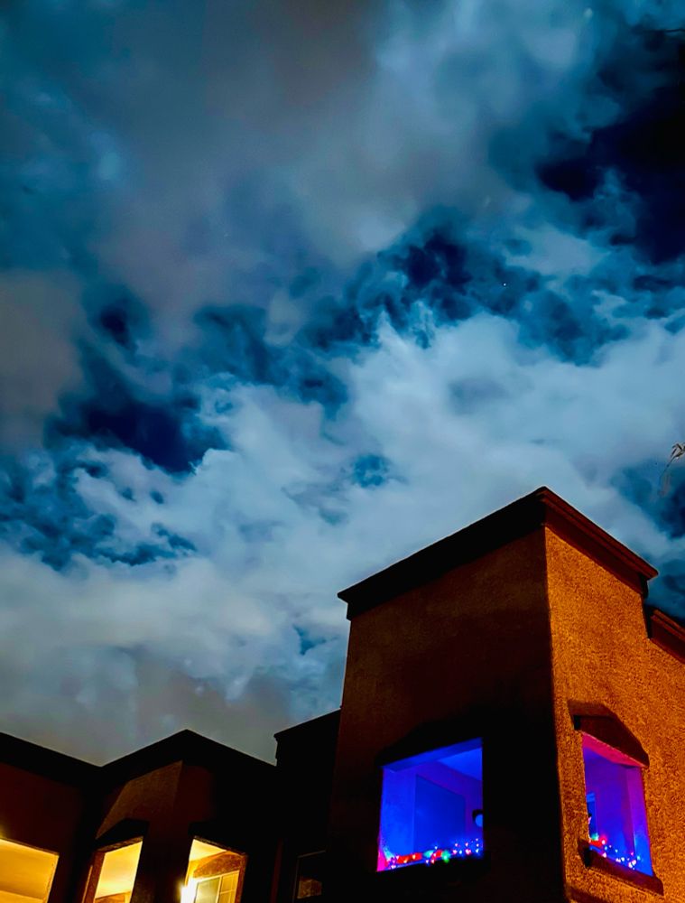 A southwestern style building with multicolor Christmas lights is backlit by the bright night sky. The clouds obscure the full moon.