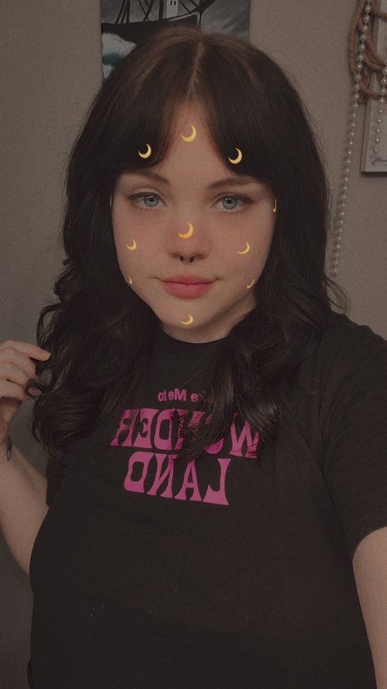 Ashe in a nice selfie with a face filter that puts small, yellow crescent moons all over her face. Her black shirt has pink lettering on it that says "Take me to Wonder Land."