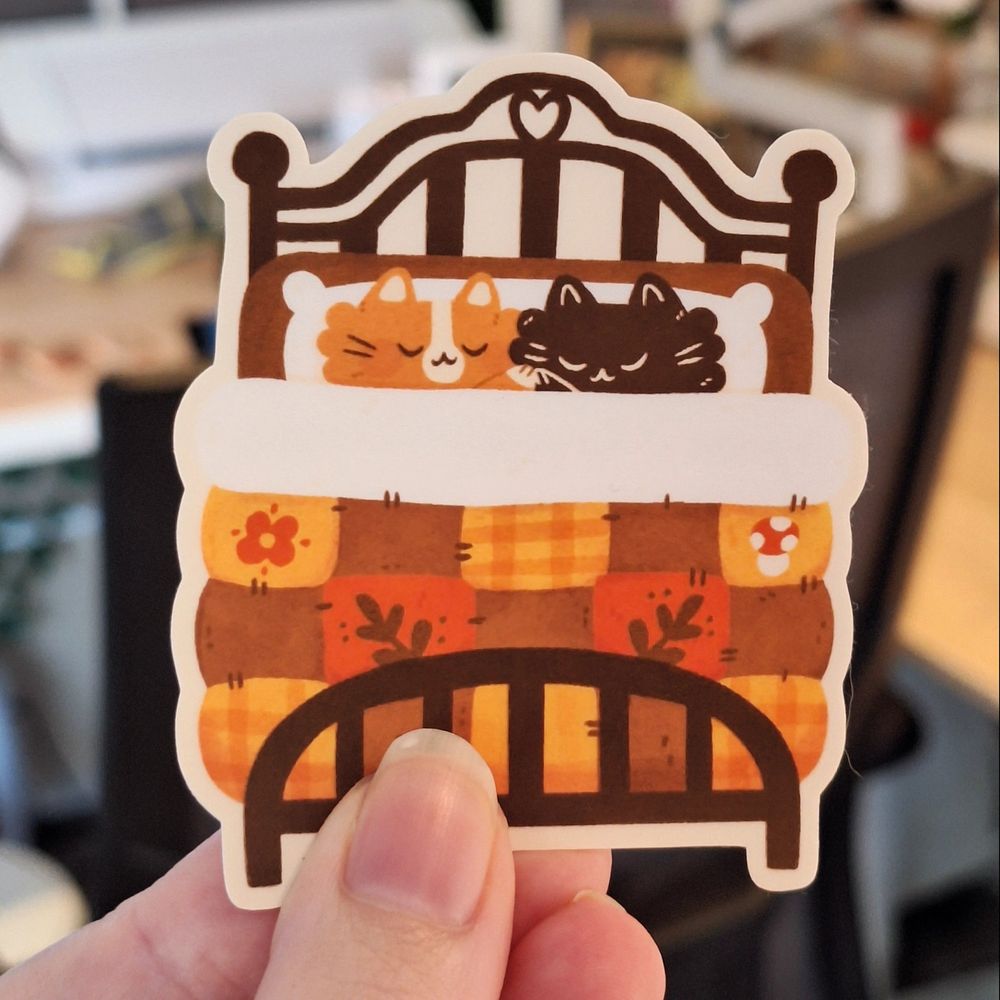 A photo of a die-cut sticker: a bed with a black cat and an orange cat holding paws. They lay under a quilted blanket