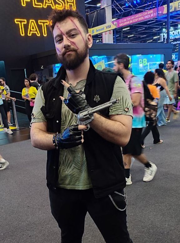 Homem segurando uma arma de brinquedo, ele usa camiseta verde, colete preto e luvas sem dedo pretas, ele possui 3 citrazes falsas no rosto, cabelo curto, barba, bigodes grandes, sua calça também é preta e com 2 correntes penduradas. Eke está em um evento de cultura pop com algumas pessoas passando atrás.