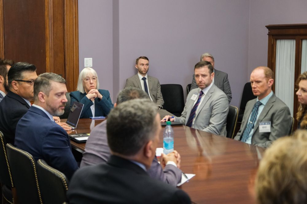 Senator Murray is sitting at the end of a large table with nine other people pictured, as well as two people on chairs against a wall. She is sitting with her hands together under her chin, looking at someone toward the end of the table with their back to the camera.