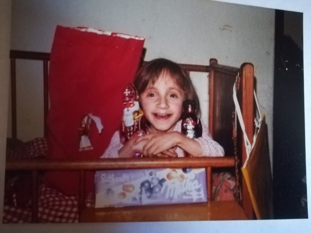 Ich in einem braunen Kinderbett, neben mir ein großer roter geöffneter Sack. In meinen Händen ein Schoko-Nikolo und -Krampus und vor mir eine sehr große Tafel Milkaschokolade. Ich strahle in die Kamera.