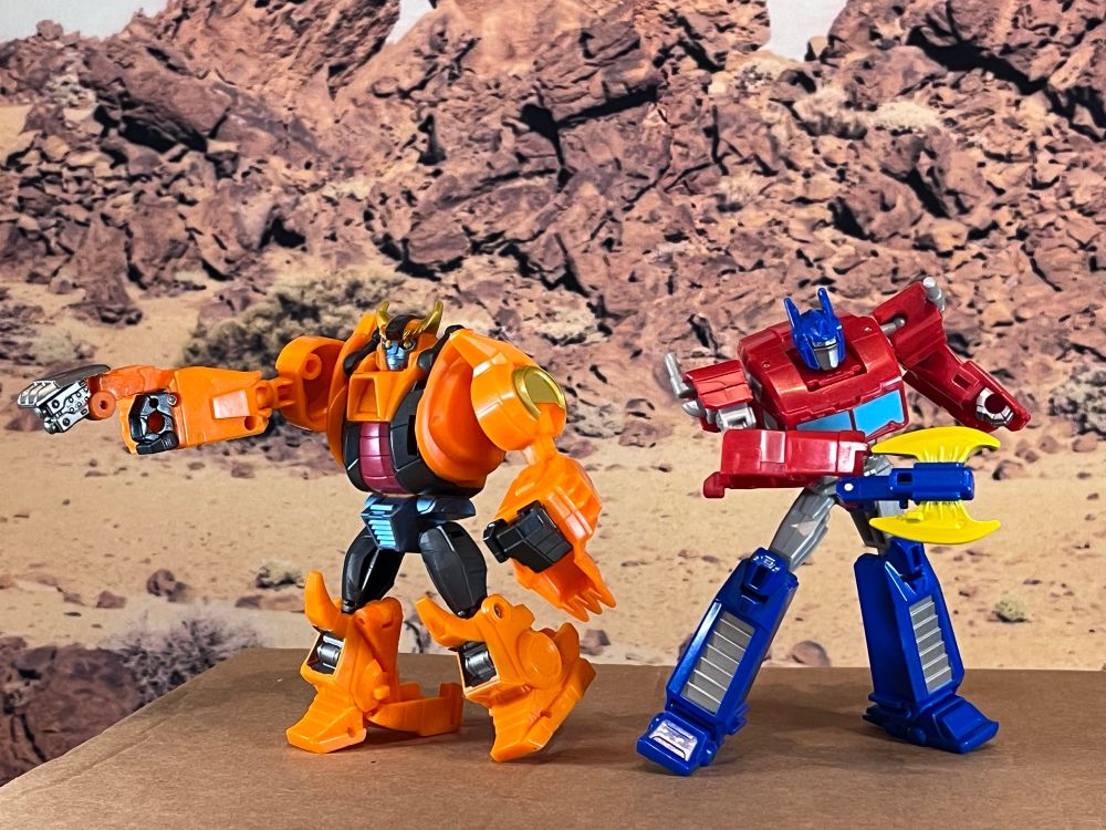 Earthspark Jawbreaker and Earthspark Optimus Prime in Robot mode. Jawbreaker is an orange chunky boy with gold horns and a sweet character. Optimus Prime is the classic red and blue truck hero and includes a blue fold out energon axe