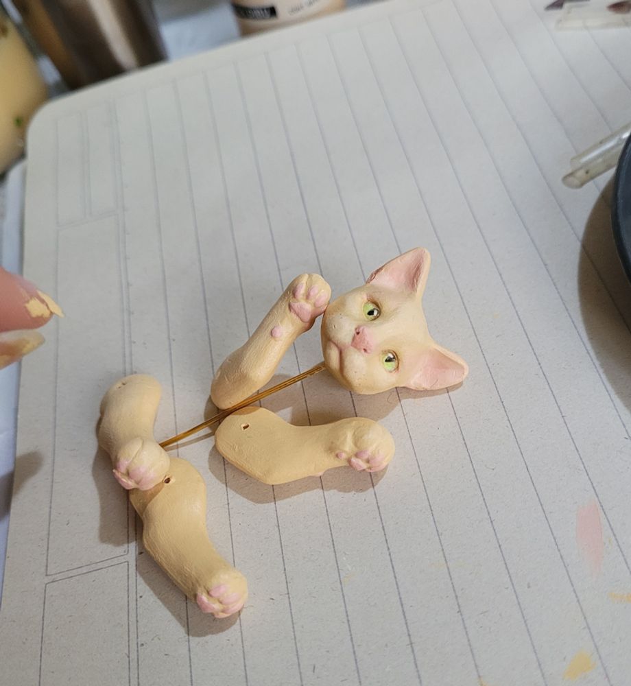 Pic of parts of a cat (made of clay). There's a head with the four paws/legs. The cat is a pale yellowish color with pink ears, nose and beans and has green eyes