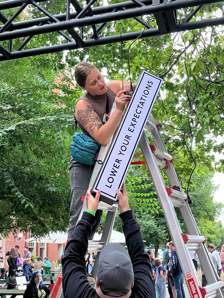A woman on a ladder lowers a black on white sign that reads "Lower Your Expectations" while a man reaches up to take it. 