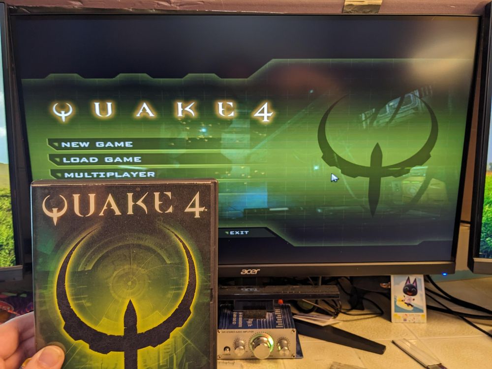 Quake 4's main menu being displayed on a modern monitor, with the box from the original physical release of the game being held up in the foreground.