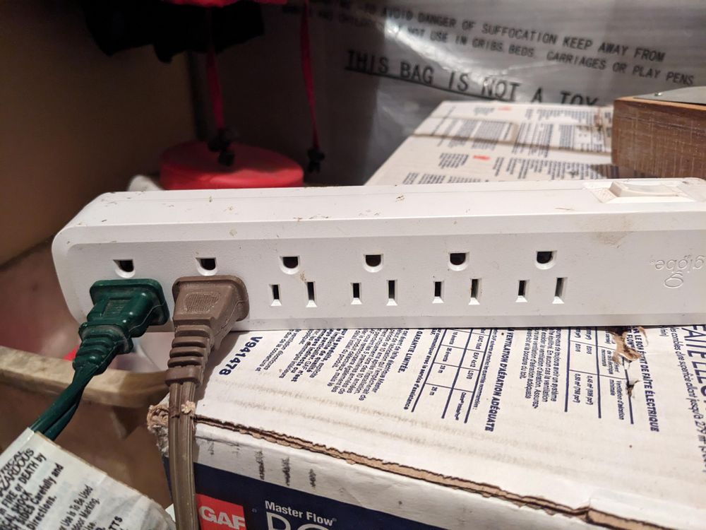 A power strip with North American-style outlets laying atop a cardboard box. Two plugs, one green and one brown, are plugged into the outlets at the end of the strip, leaving four unused.