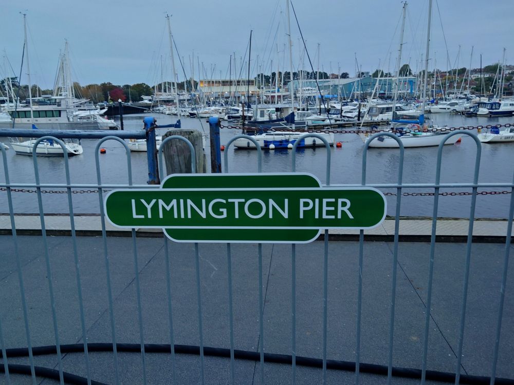 Station sign on railings at Lymington Pier station with Lymington River behind and boats moored.