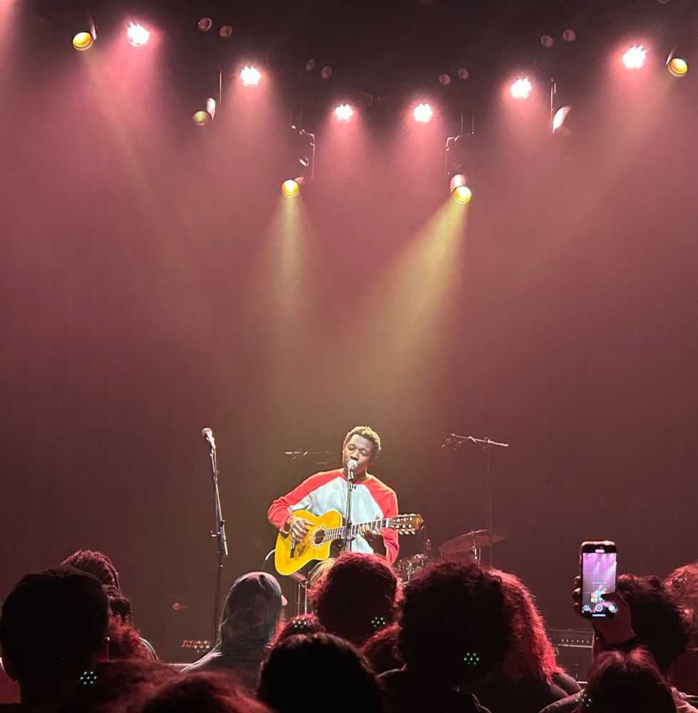 John Roseboro seated on stage at a mic with a guitar, crowd in foreground.