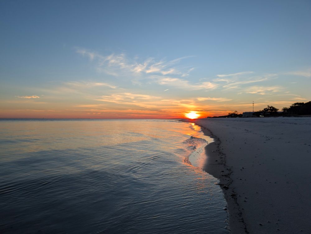 The sun setting over the beach, the shore still wet from the last wave reflects the orange, blue, and pink of the sky around the setting sun.