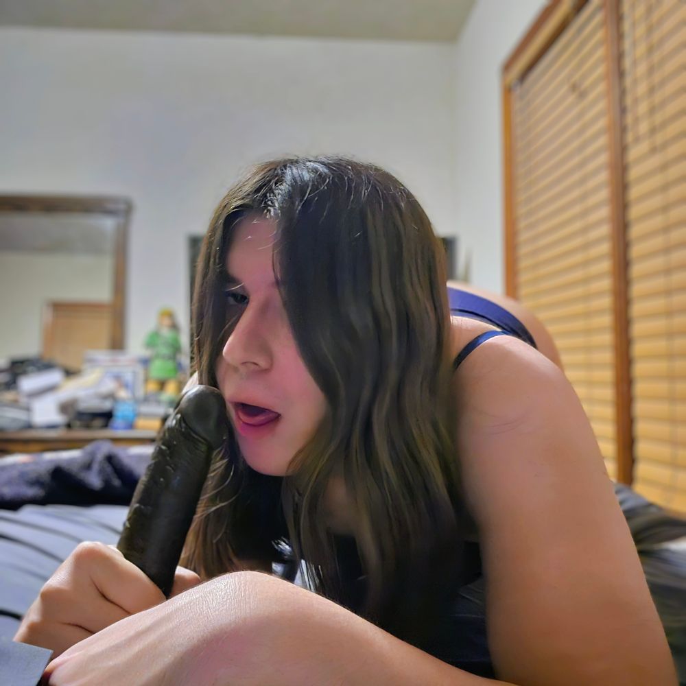 Isabella with the dildo next to her mouth with her tongue slightly out while wearing a blue bra and a pair of blue panties.
