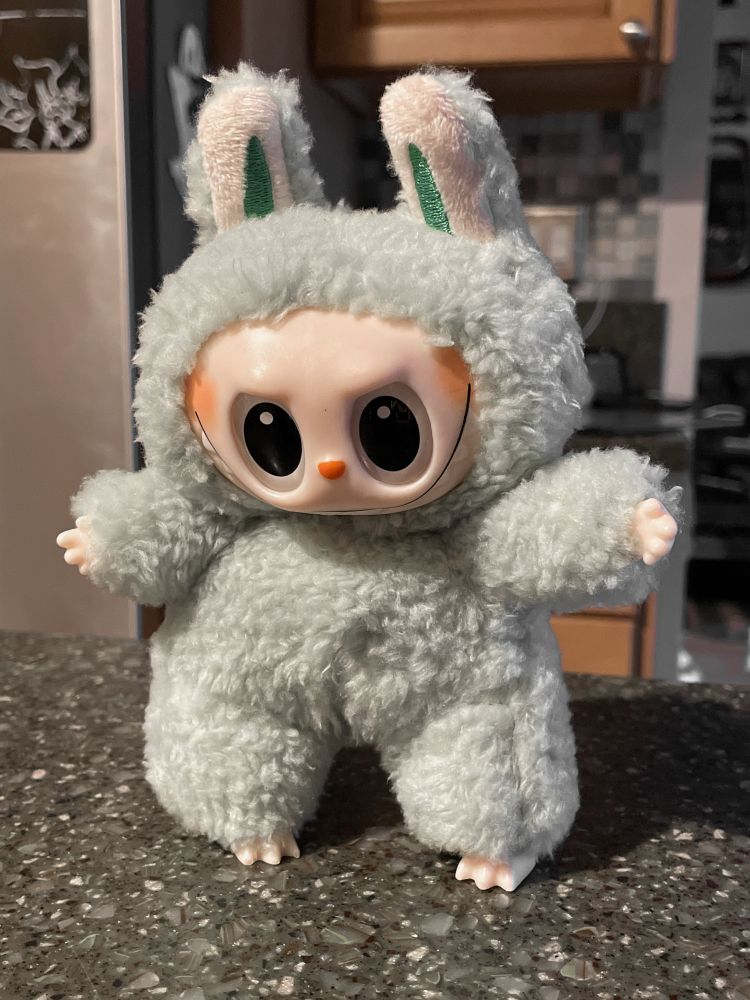 A knockoff Labubu is standing on a counter. One arm is smaller than the other