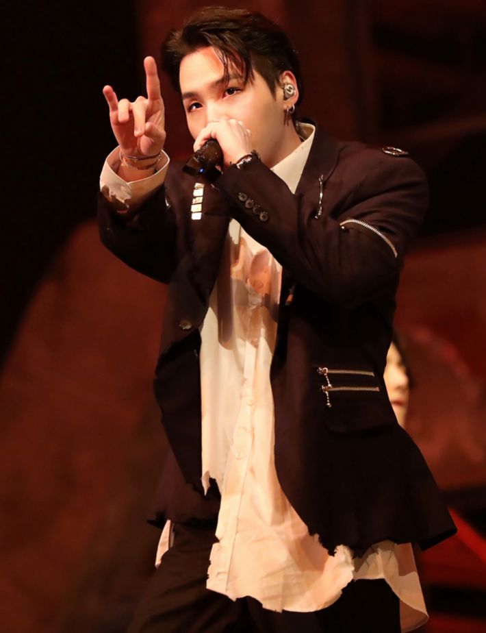 Min Yoongi from the Map of the Soul ON:E virtual concert in 2020. He has black hair and an undercut with his bangs swept  back and to the side. She’s wearing a school uniform or suit of white button up and black jacket and pants that have been deconstructed and punk-ified with strategic rips and safety pins and zippers. He’s singing into a black mic that he’s holding up to his face and his other arm is extended out in front of him, pointing with his fingers in a formation reminiscent of devil horns 