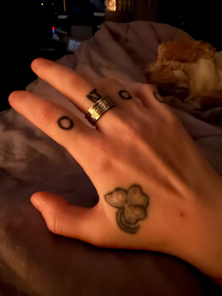 A photograph of a right hand. A black and gray shamrock is tattooed on it. The knuckles are tattooed to say Do No. there is a silver ring on the middle finger. 