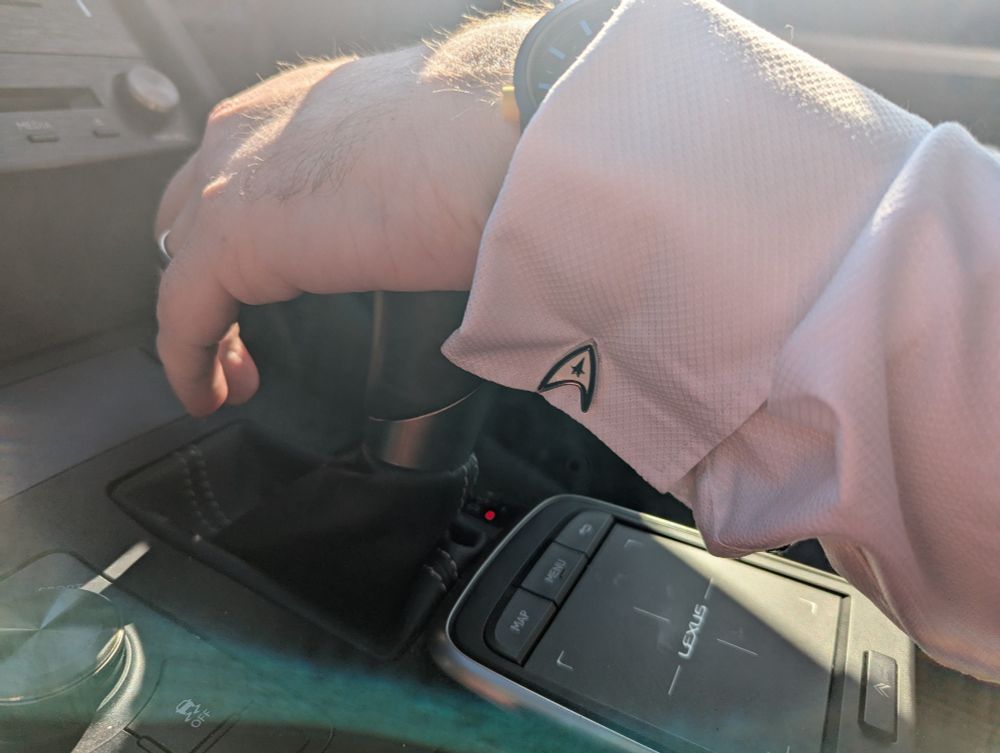 A man's hand on the gear shifter of a car, wearing a white dress shirt with Starfleet delta cufflinks.