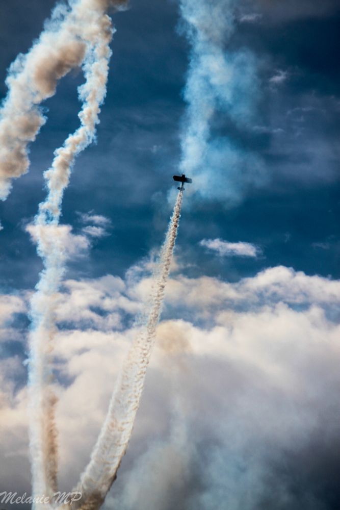 Ein Farbfoto im Hochformat. Ein Kunstflug mit einem kleinen Flugzeug das Qualm hinter sich her zieht und gerade von unten nach oben fliegt am wolkig blauem Himmel 