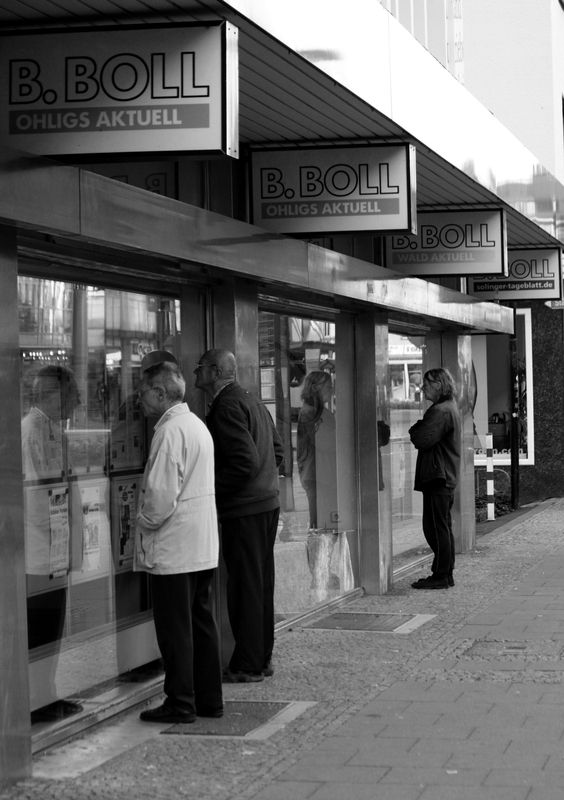 Ein Schwarz-Weißfoto im Hochformat. Drei Männer stehen nah vor einem Schaufenster einer Tageszeitung und lesen 